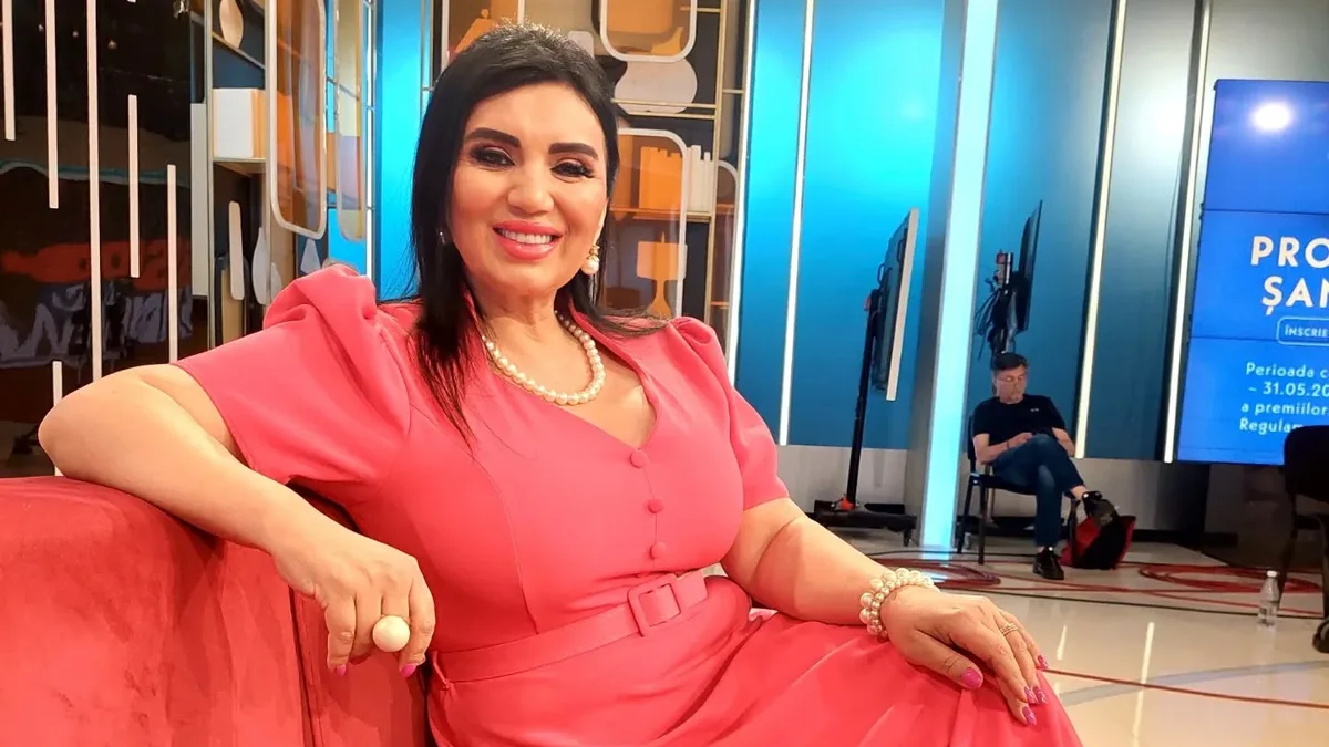 Adriana Bahmuțeanu, în rochie roz și cu un colier de perle, zâmbește de pe o canapea roșie într-un studio TV.