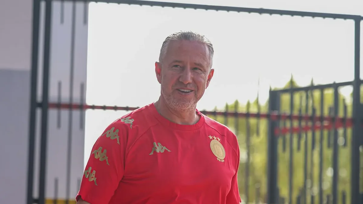 Laurențiu Reghecampf în echipament sportiv roșu, în aer liber, lângă un gard metalic.