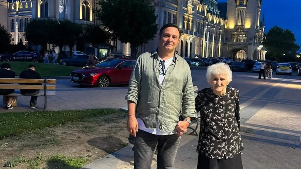 Un bărbat și o femeie în vârstă se țin de mână, seara, în fața unei clădiri monumentale iluminate.
