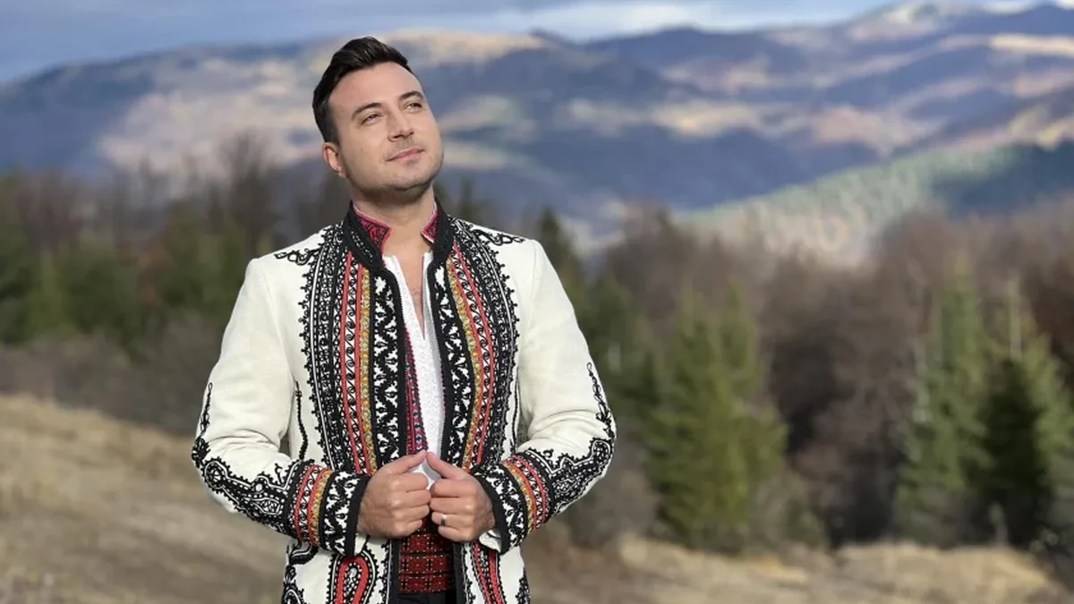 Valentin Sanfira în costum popular brodat, în fața unui peisaj cu păduri și munți.