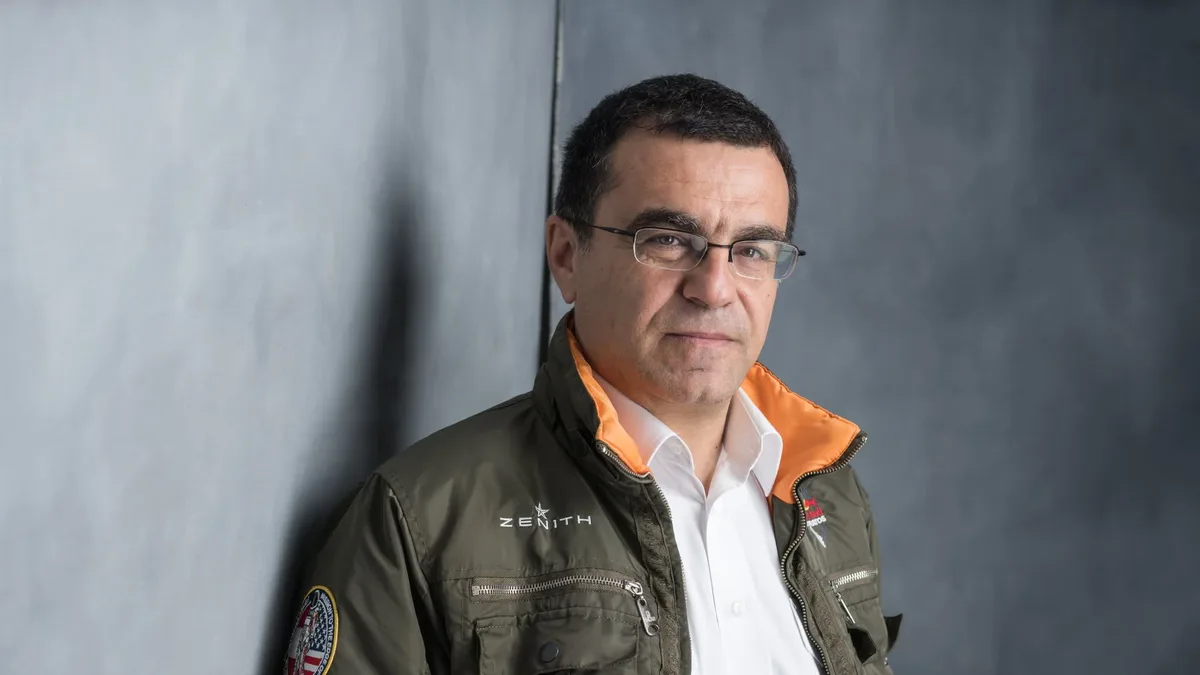 Mihai Mărgineanu, cu ochelari, cămașă albă și jachetă verde cu guler portocaliu, lângă un perete gri.