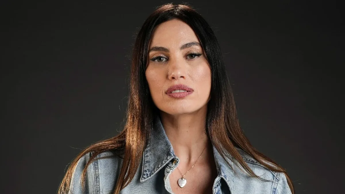 Alina Pușcău poartă o cămașă din denim și un colier cu pandantiv în formă de inimă, pe un fundal închis.
